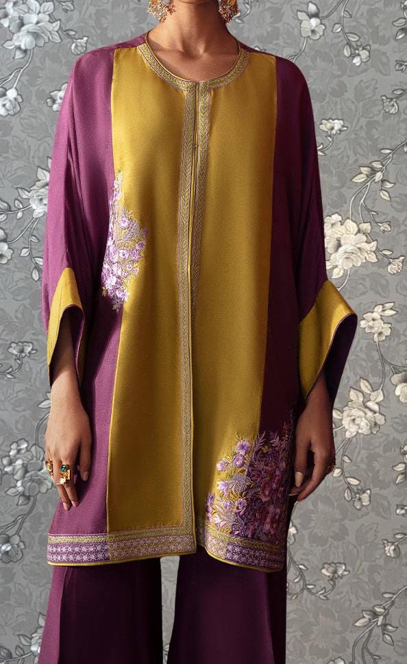 A person wearing a colorful, patterned dress with a yellow and purple color scheme, standing against a floral background.