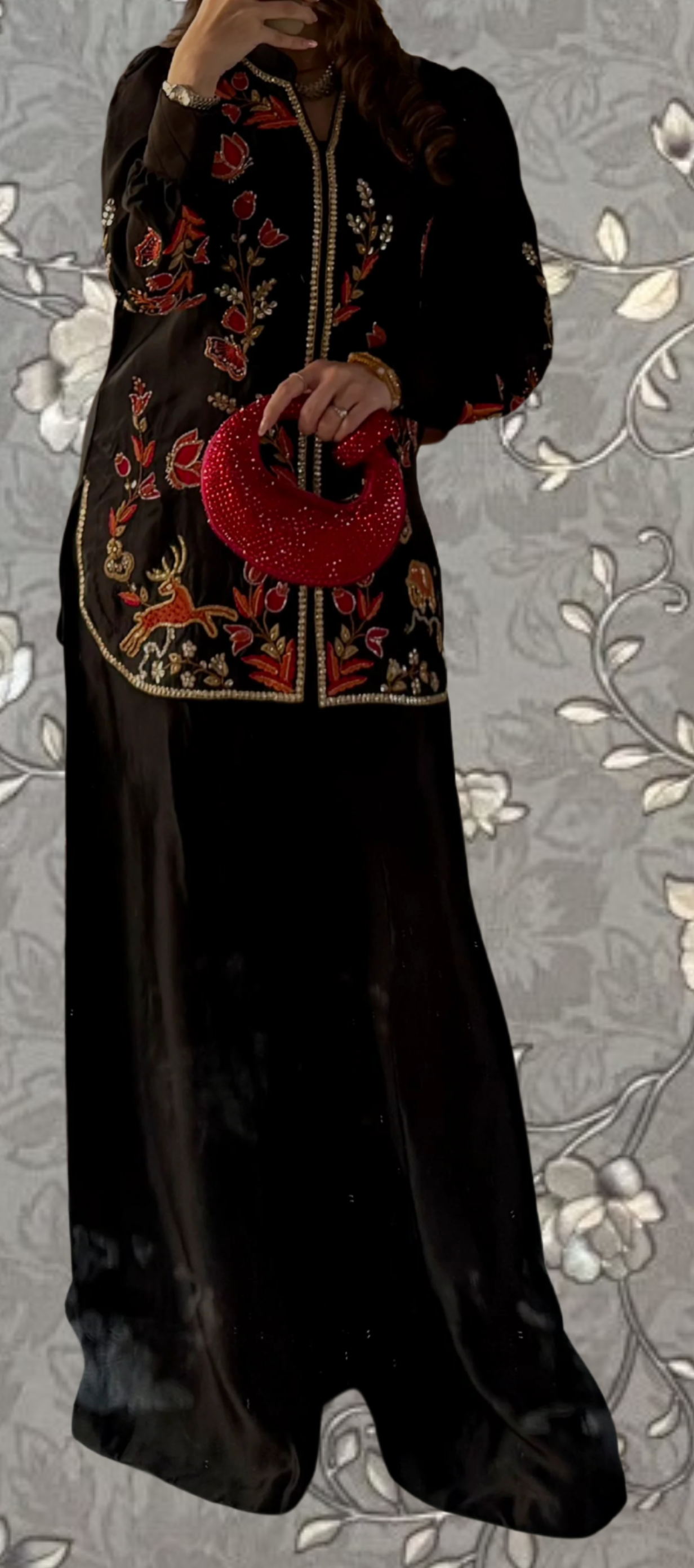 A woman wearing a long black dress with a floral pattern and a red purse is standing against a gray floral background.