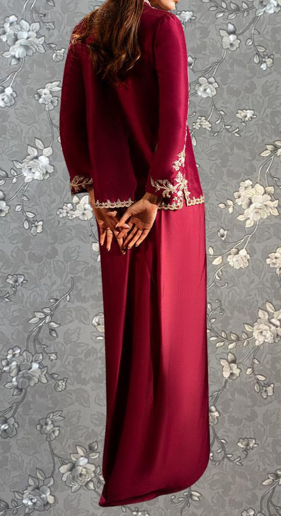 A woman wearing a long, burgundy dress with intricate embroidery stands against a gray floral background.