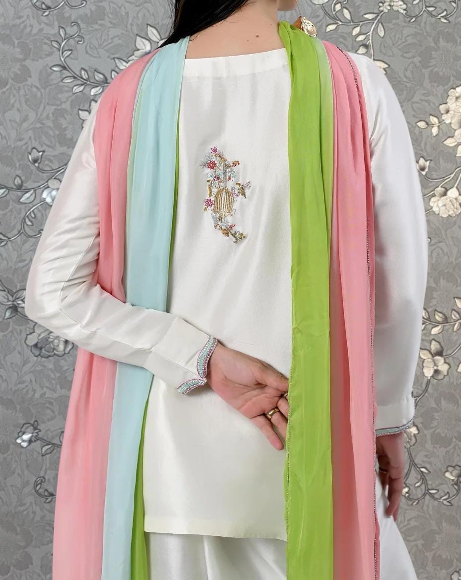 A person wearing a white dress with a colorful scarf draped over their shoulders, standing against a gray floral background.
