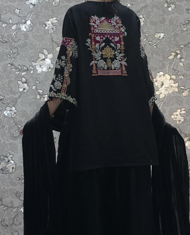 A person wearing a black dress with intricate embroidered designs on the sleeves and back, standing against a floral patterned background.