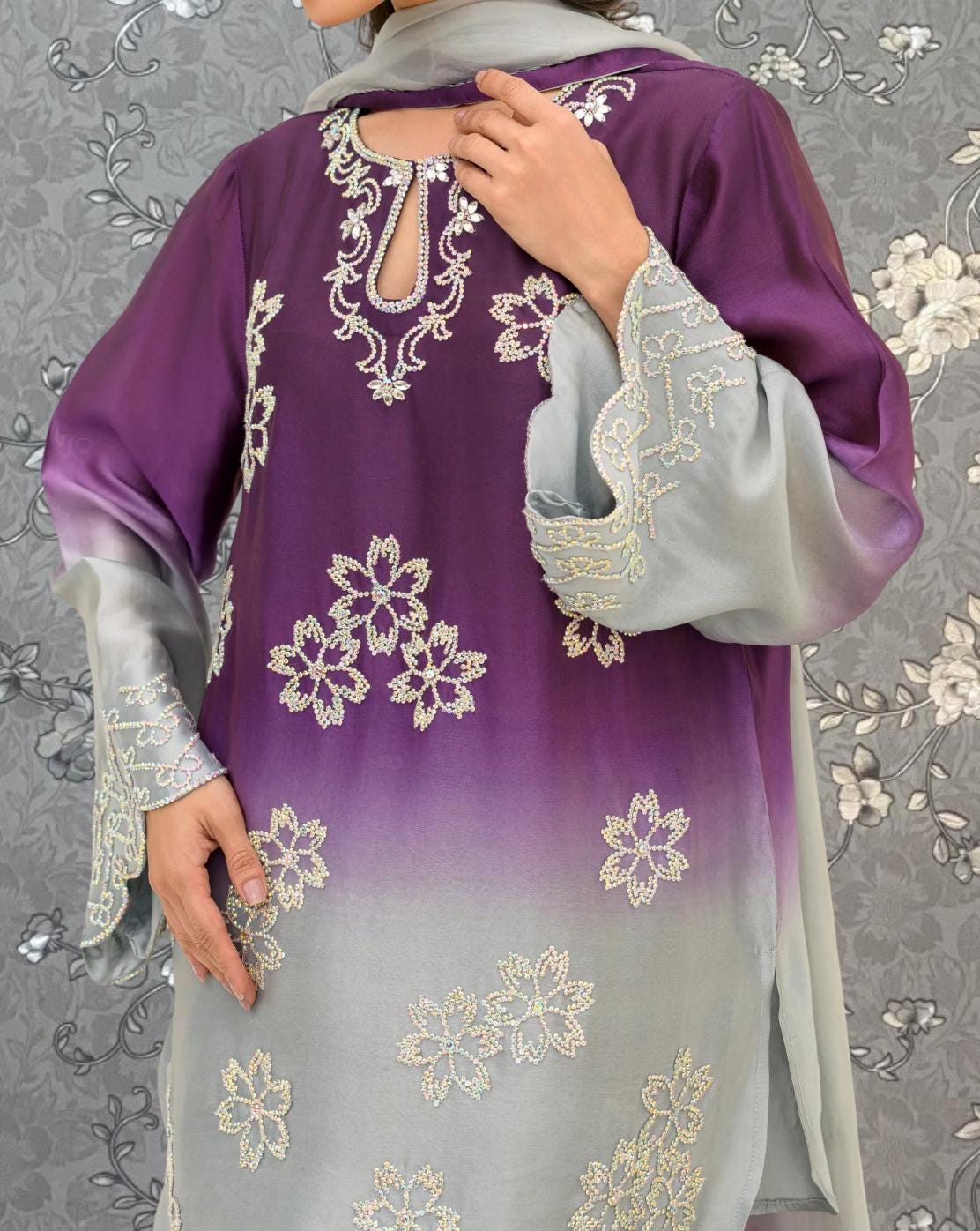 A woman wearing a purple and gray dress with intricate embroidered designs, including flowers and floral patterns, stands against a gray floral background.