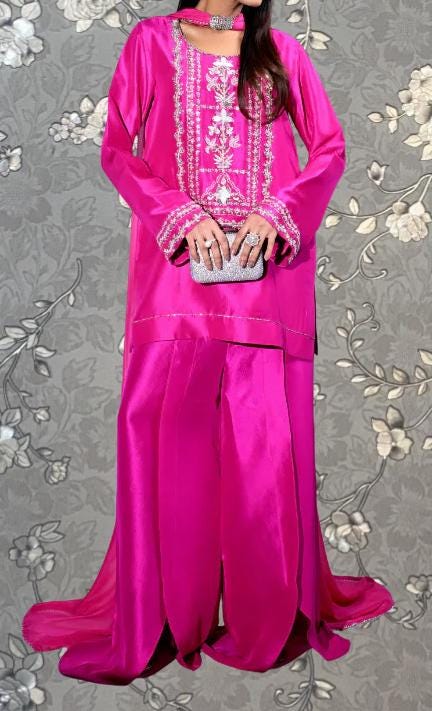 A woman wearing a long pink dress with intricate embroidery and a matching pink skirt, holding a small clutch purse.