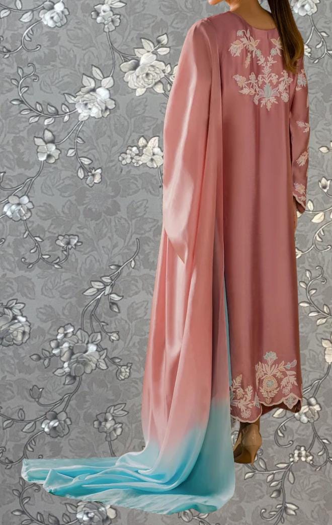 A woman wearing a pink dress with floral embroidery stands against a gray floral background.