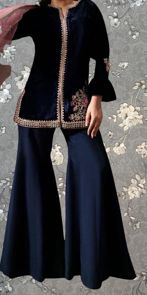 A woman wearing a black velvet dress with gold embroidery and a matching black skirt, posing against a floral background.