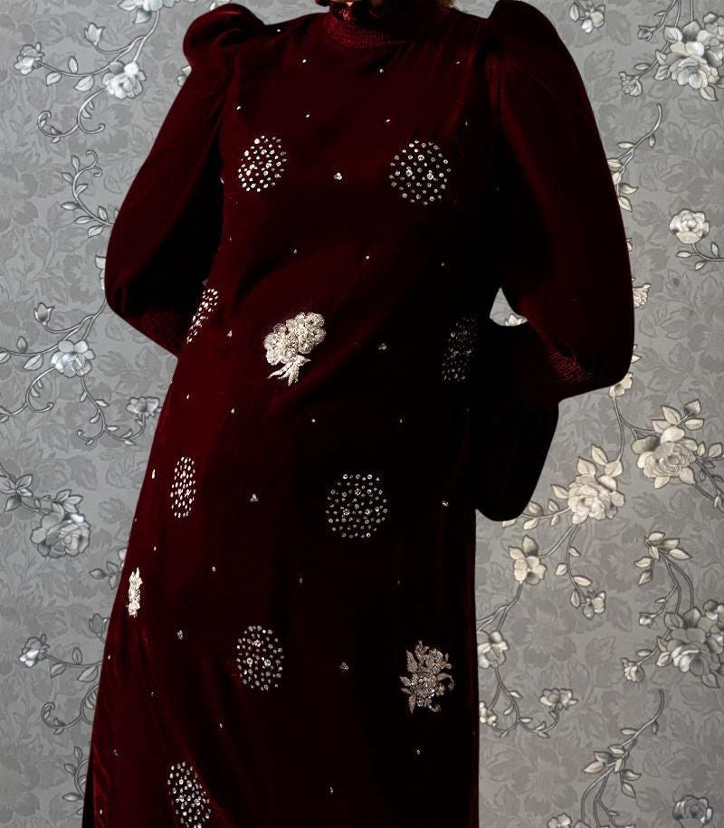 A close-up of a burgundy dress with white floral embroidery on the bodice and sleeves, standing against a gray floral background.