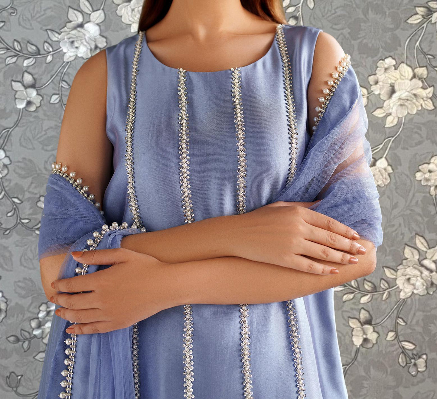 A woman wearing a blue dress with silver embellishments stands with her arms crossed against a gray floral background.