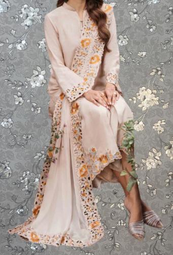 A woman wearing a long, light pink dress with floral embroidery, sitting against a gray floral background.