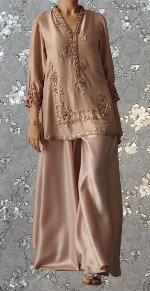 A woman wearing a long, pink dress with intricate embroidery and a high collar, standing against a gray floral background.