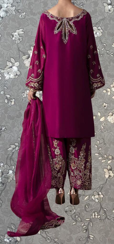 A person wearing a vibrant purple dress with intricate embroidery and a matching purple skirt, standing against a gray floral background.