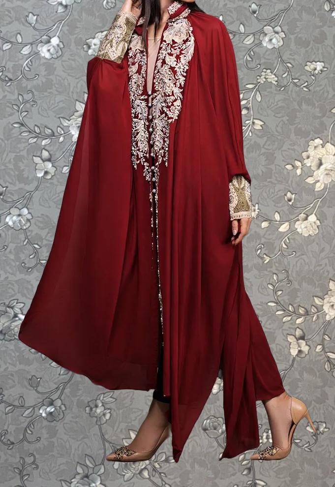 A person wearing a long, red cape-like garment with intricate embroidery and gold accents, standing against a gray floral background.