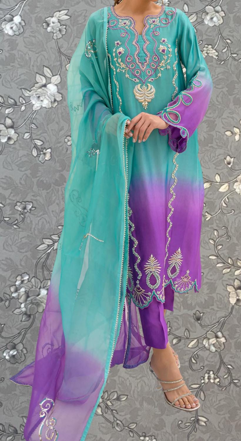 A person wearing a colorful, patterned dress with a turquoise and purple color scheme, standing against a gray floral background.