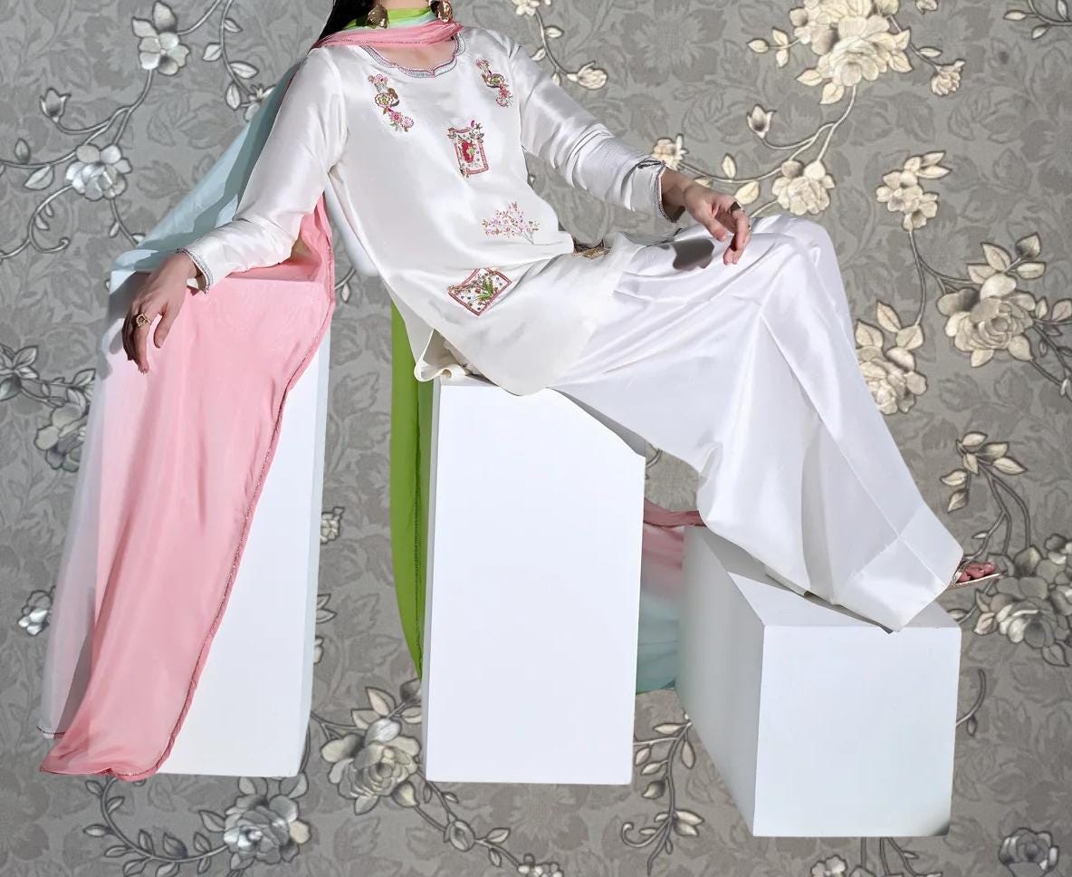 A person wearing a white dress with pink accents is sitting on a white pedestal against a gray floral background.
