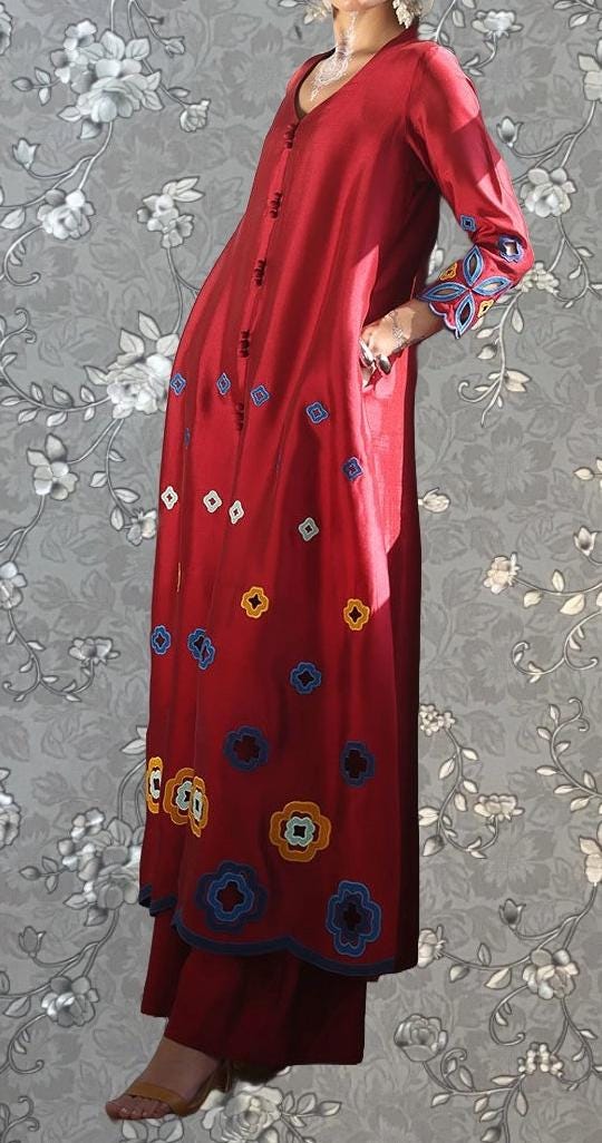 A woman wearing a long red dress with colorful floral embroidery stands against a gray floral background.