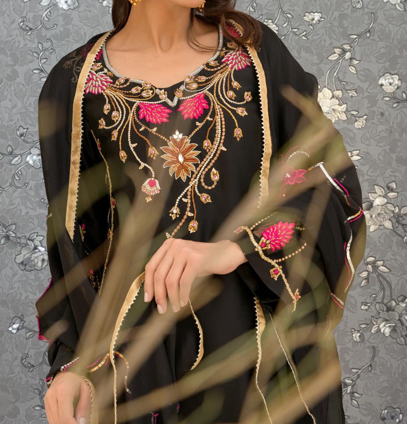 A woman wearing a black dress with intricate gold embroidery and pink accents, posing against a gray floral background.