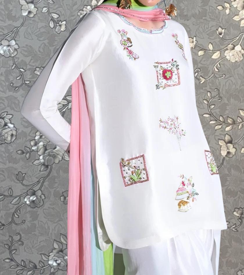 A person wearing a white dress with pink and green accents, featuring floral embroidery and a pink scarf draped over the shoulder.