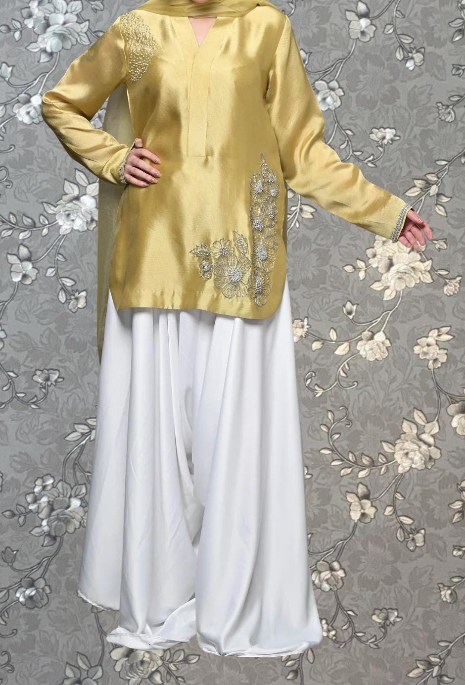 A woman wearing a gold and white dress with a floral pattern on the top and skirt, standing against a gray floral background.
