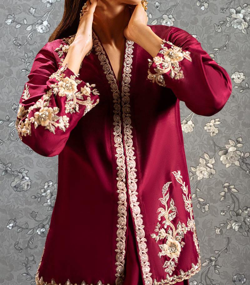 A woman wearing a red and gold embroidered dress stands in front of a gray floral patterned background.