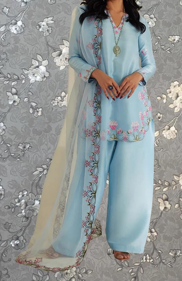 A woman wearing a light blue dress with floral embroidery and a matching shawl stands against a gray floral background.