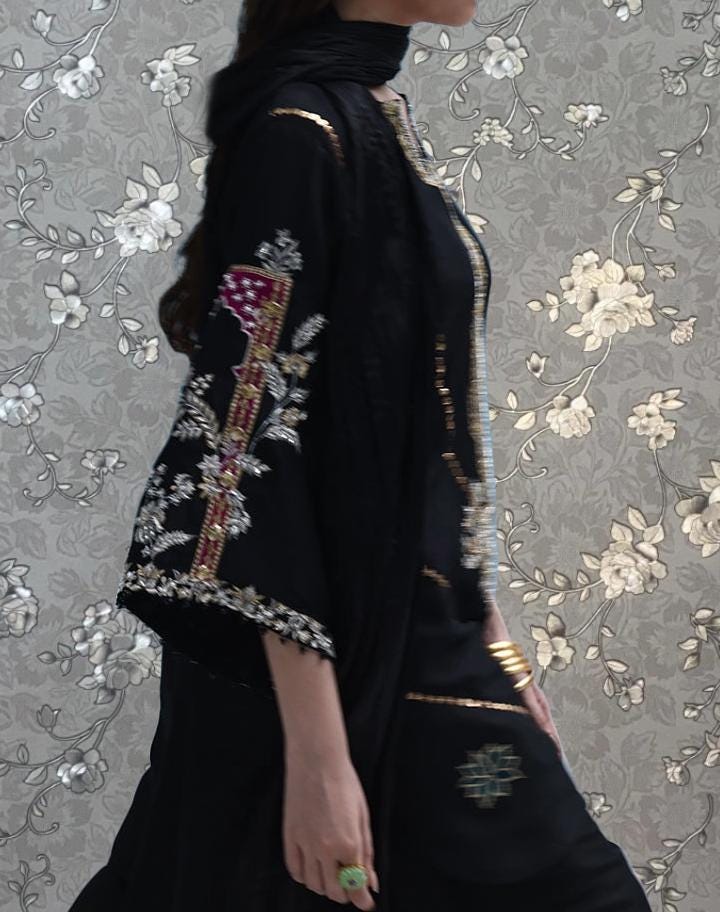 A woman wearing a black dress with intricate embroidery and floral patterns, standing against a floral patterned background.