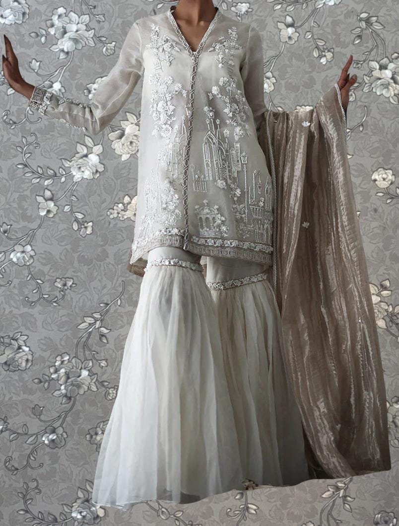 A woman wearing a white dress with intricate embroidery stands against a gray floral background.