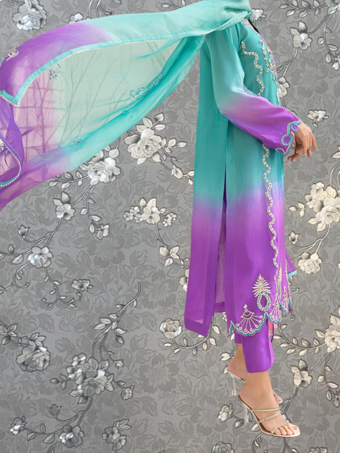 A person wearing a long purple and teal dress with floral embroidery stands against a gray floral background.