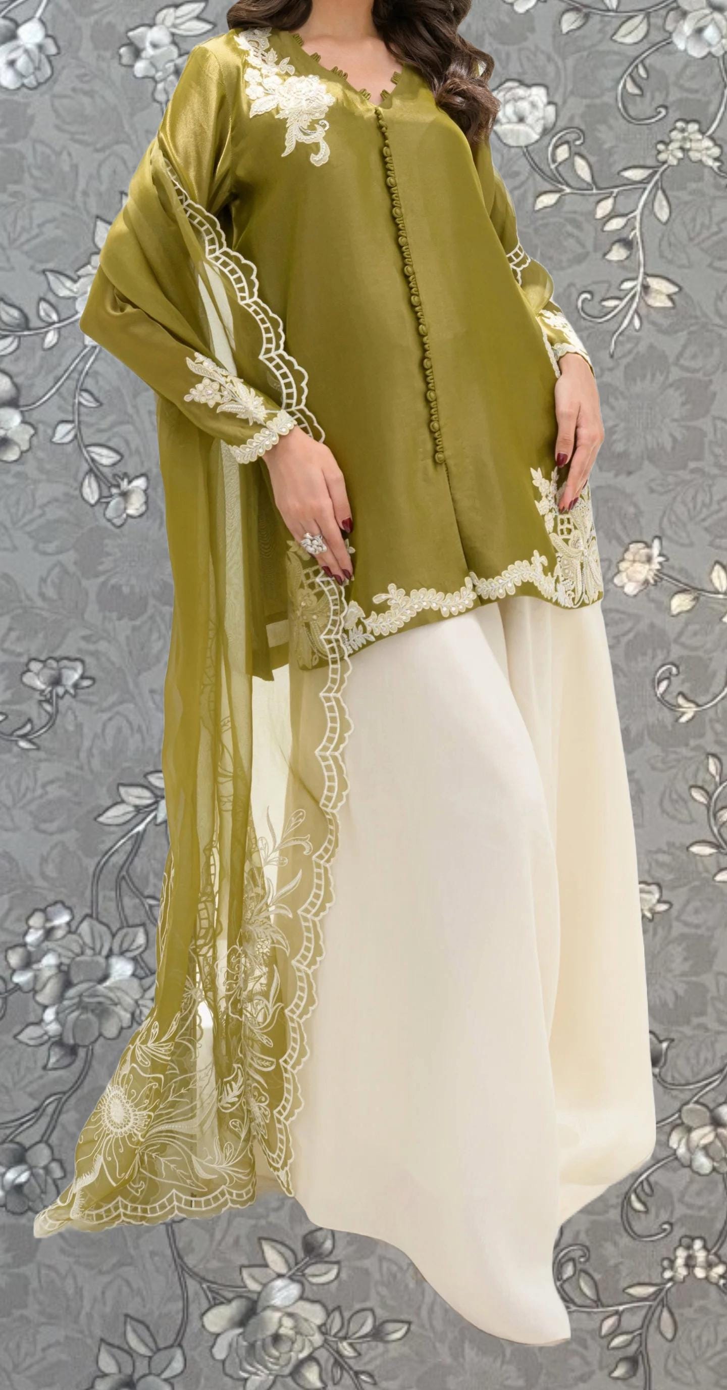 A woman wearing a green and white dress with intricate embroidery and lace details, standing against a gray floral background.