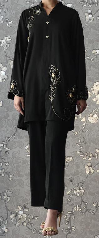A person wearing a black outfit with gold embroidered designs on the sleeves and pants, standing against a gray floral background.