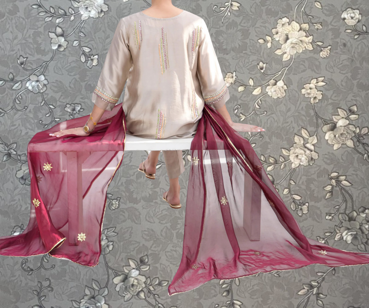 A person sitting on a bench wearing a white dress with colorful embroidery and a long red shawl.