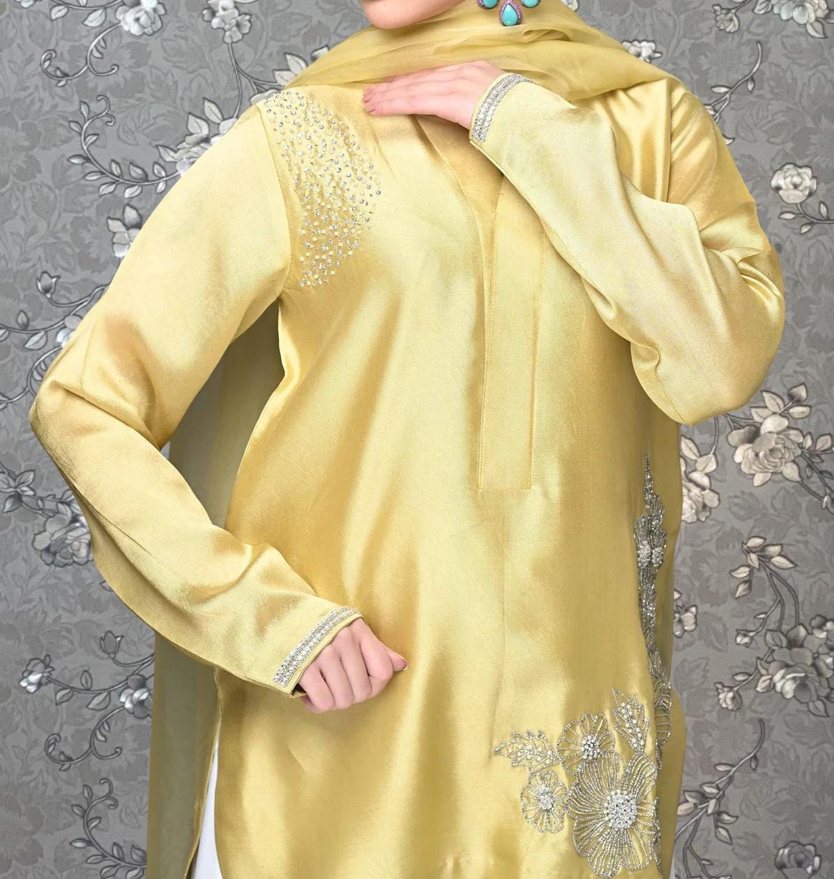 A person wearing a yellow dress with silver embroidery and a matching yellow scarf stands against a gray floral background.