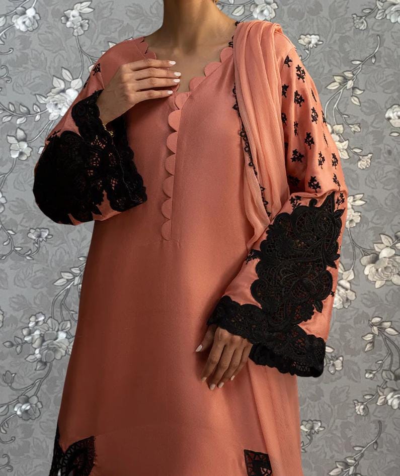 A woman wearing a peach-colored dress with black lace accents, posing against a gray floral background.