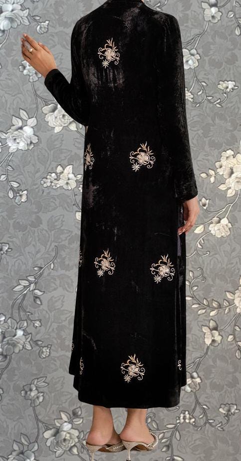 A person wearing a long black velvet dress with gold floral embroidery stands against a gray floral background.