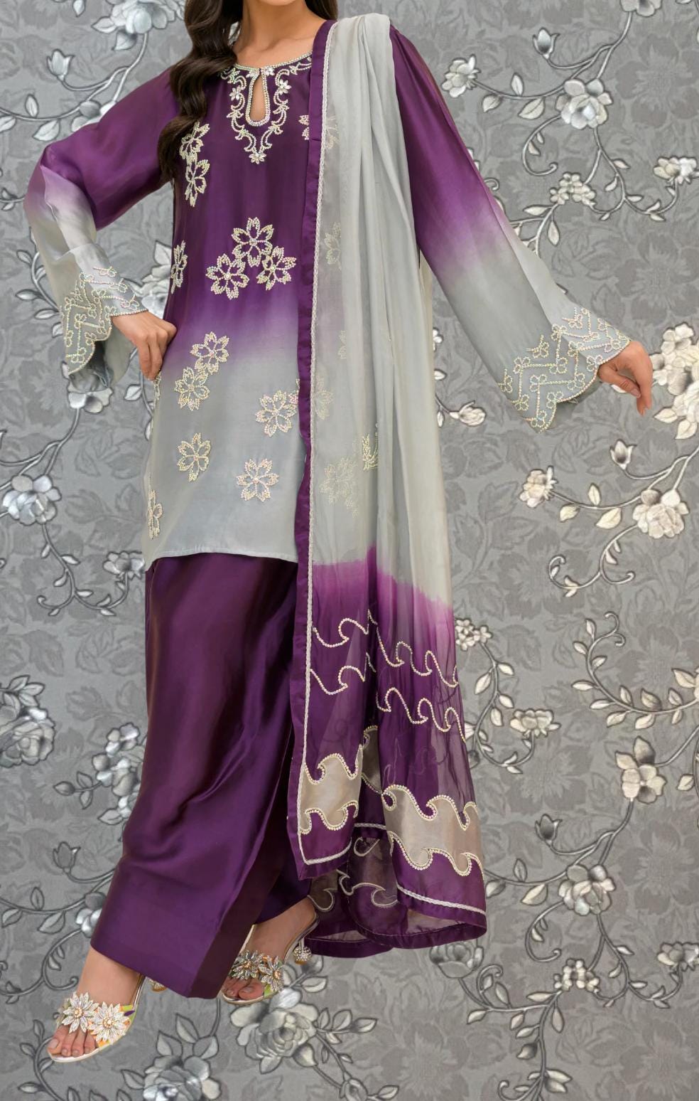 A woman wearing a purple and white dress with a gray shawl, standing against a gray floral background.