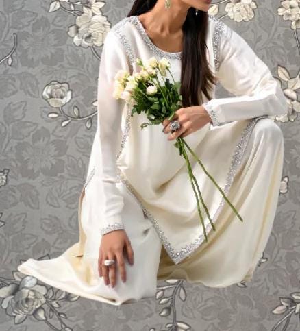 A woman in a white dress holding a bouquet of white flowers, kneeling against a gray floral background.