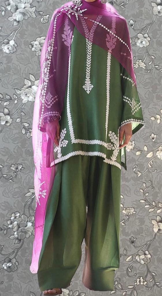 A person wearing a green and pink dress with white embroidered patterns, standing against a gray floral background.