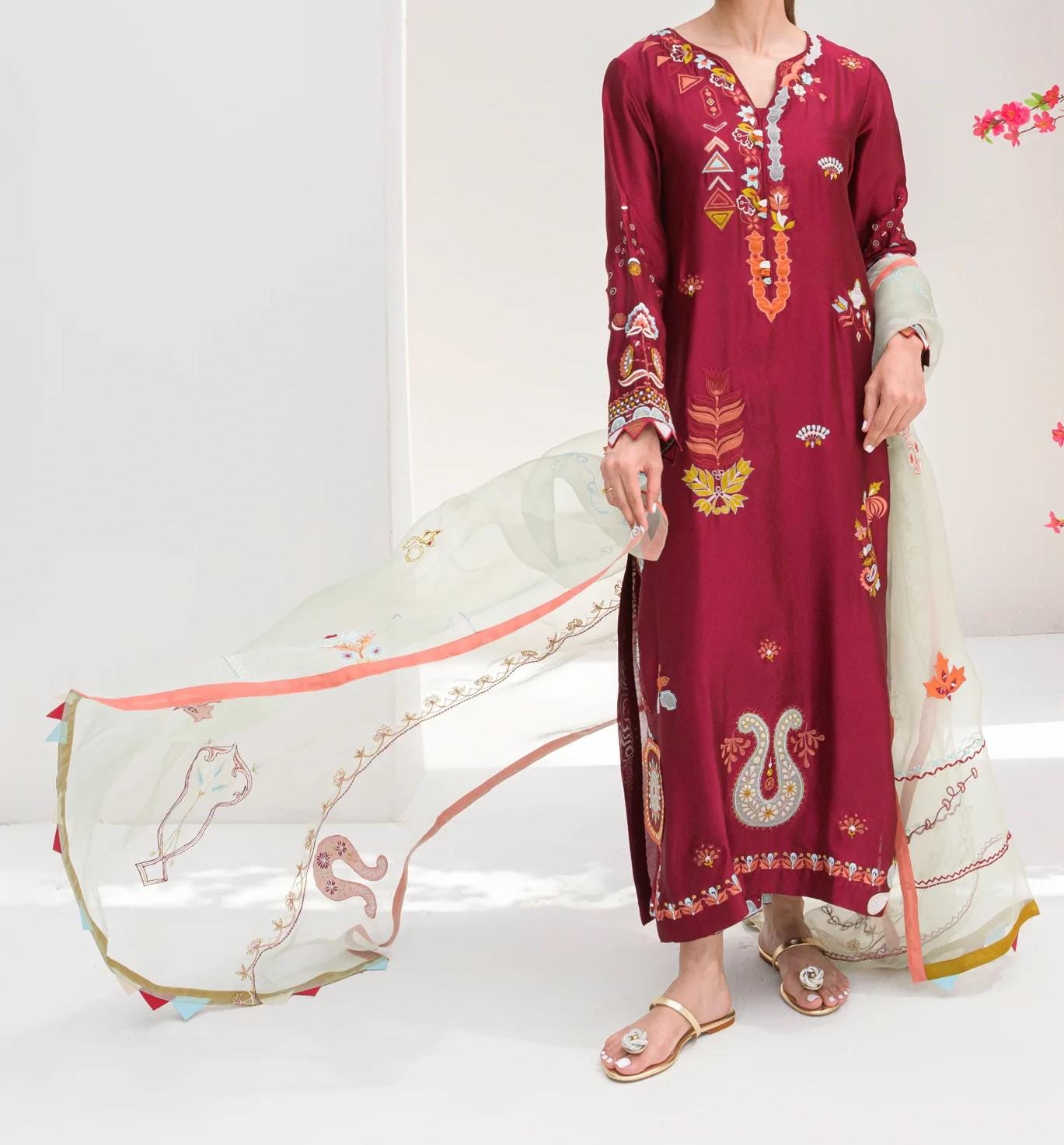 A person wearing a red and gold embroidered dress stands in front of a white background, holding a long white fabric with a floral pattern.
