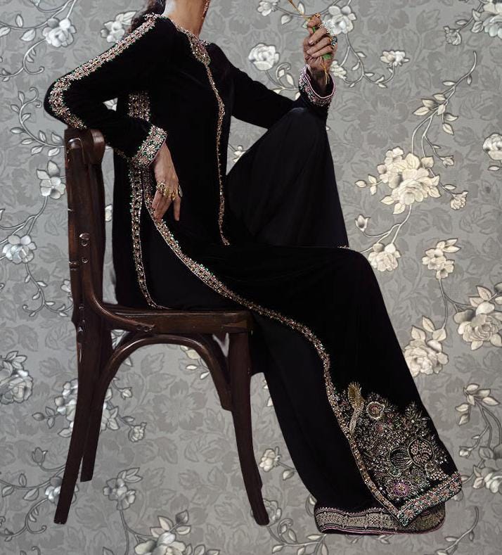 A woman wearing a long black dress with intricate embroidery is sitting on a wooden chair against a floral patterned background.