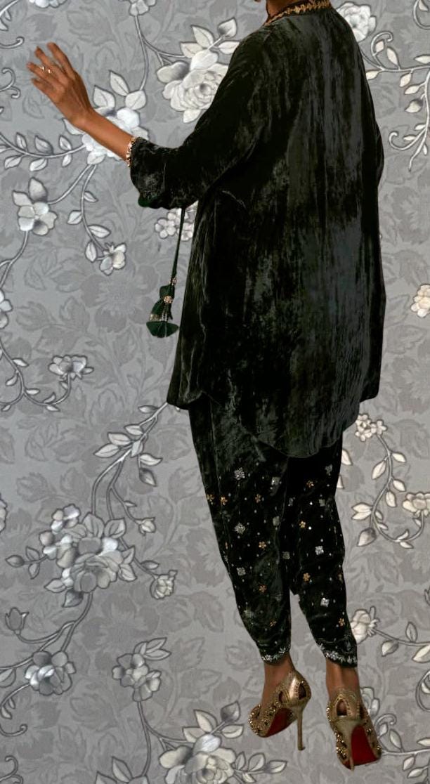 A person wearing a black velvet outfit with gold accents and high heels is standing against a gray floral background.