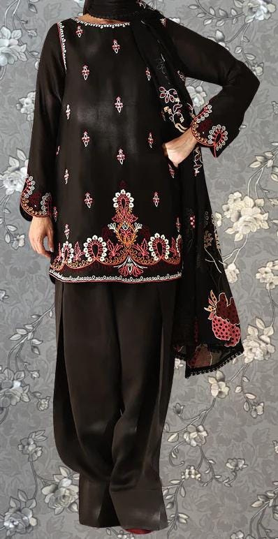 A person wearing a black dress with intricate embroidered patterns and designs, standing against a gray floral background.