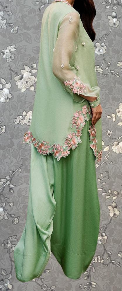 A woman wearing a green dress with pink floral accents, standing against a gray floral background.