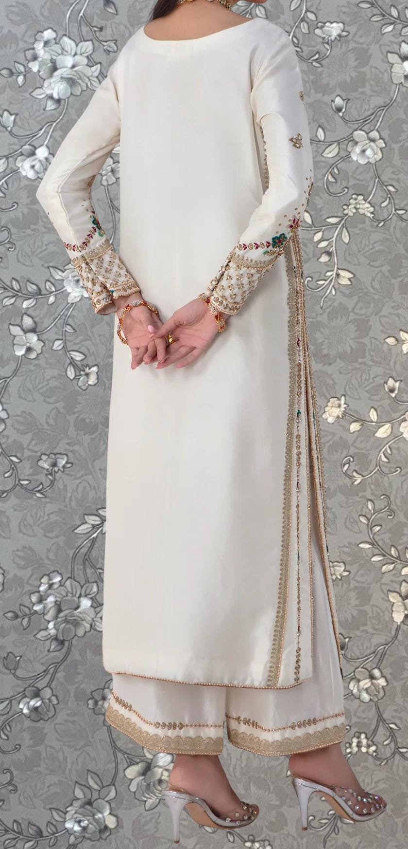 A person wearing a white dress with gold embroidery and jewelry, standing against a gray floral background.