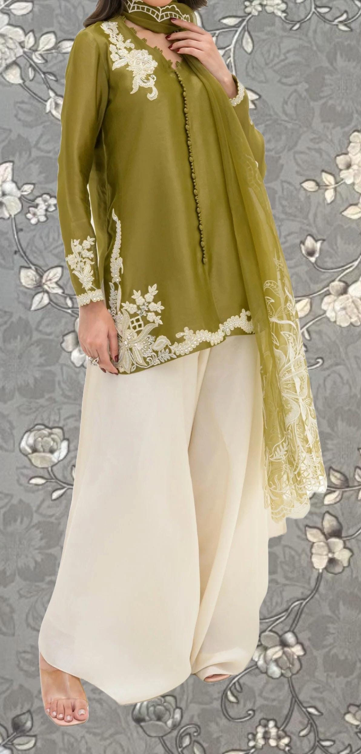 A woman wearing a green and white dress with intricate embroidery, standing against a gray floral background.