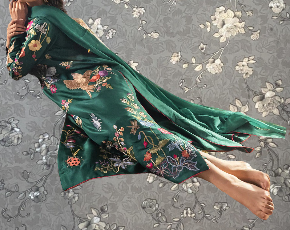 A woman wearing a green dress with intricate floral embroidery is shown in mid-air against a gray floral background.