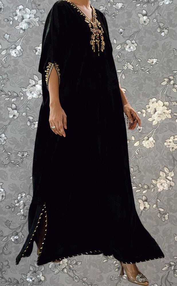 A person wearing a long black dress with gold accents and a high neckline stands against a gray floral background.