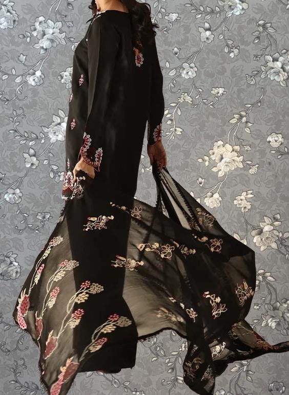 A woman wearing a long black dress with floral embroidery is standing against a gray floral background.