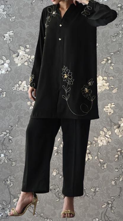 A person wearing a black outfit with gold embroidered designs, standing against a gray floral background.