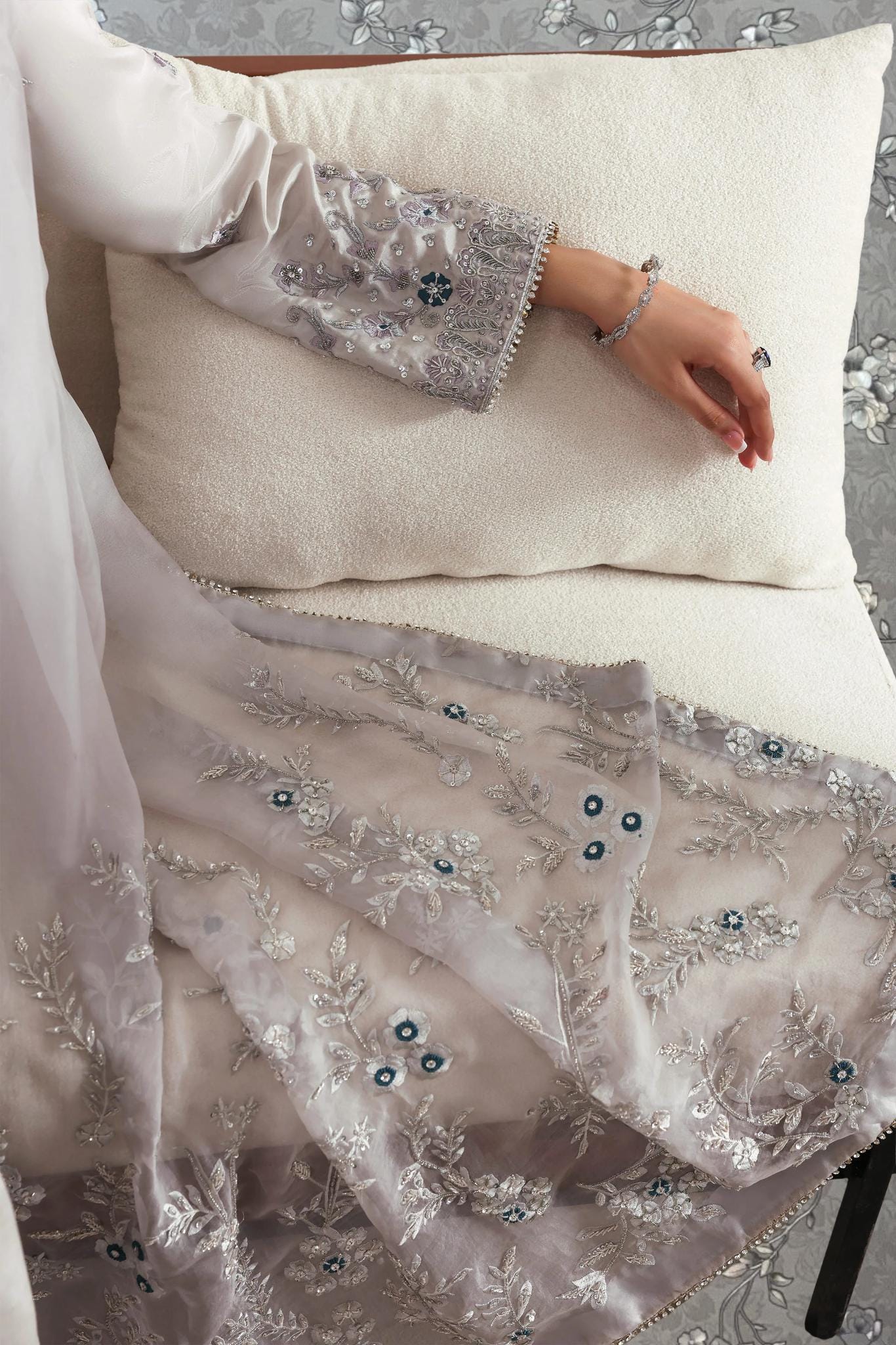 A person is lying on a bed with a white pillow, wearing a gray and white patterned dress with intricate embroidery.