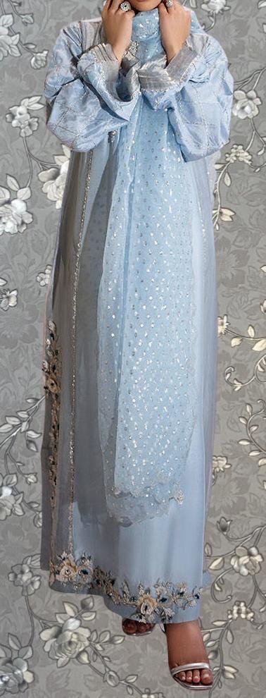 A woman wearing a long, light blue dress with a patterned scarf, standing against a gray floral background.