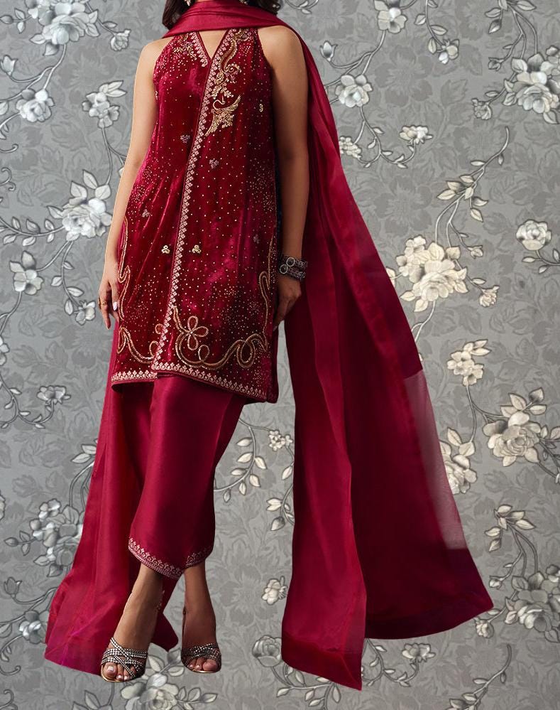 A woman wearing a red and gold embroidered dress with a matching red shawl, standing against a gray floral background.