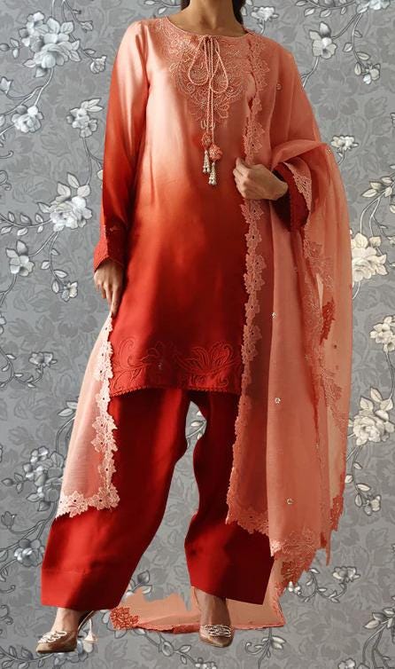 A woman wearing a vibrant red and peach-colored outfit with intricate embroidery and lace details, standing against a gray floral background.
